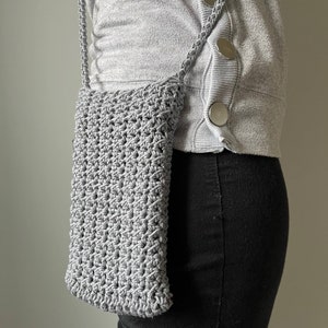 May include: A gray crocheted crossbody bag with a long strap. The bag is worn over a gray sweater and black jeans.