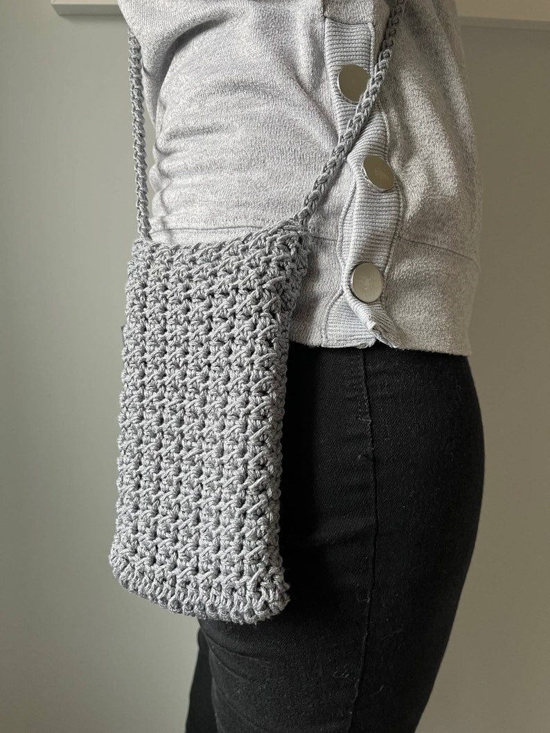 May include: A gray crocheted crossbody bag with a long strap. The bag is worn over a gray sweater and black jeans.