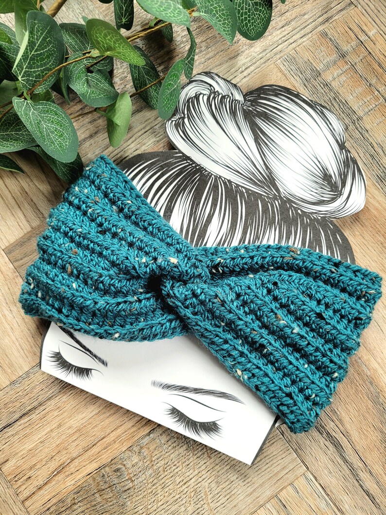 May include: A teal blue crocheted headband with a knotted design. The headband is on a white background with a drawing of a woman's face.