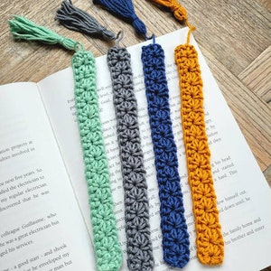 May include: Five crocheted bookmarks with tassels in different colors: green, gray, blue, and orange. The bookmarks are made with a simple stitch pattern and have a fringed tassel at the end.
