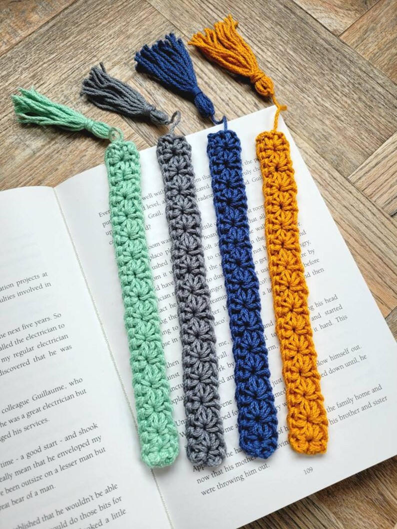 May include: Five crocheted bookmarks with tassels in different colors: green, gray, blue, and orange. The bookmarks are made with a simple stitch pattern and have a fringed tassel at the end.