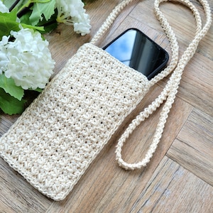 May include: A crocheted beige phone case with a long strap. The case is lying on a wooden surface with a white flower in the background.