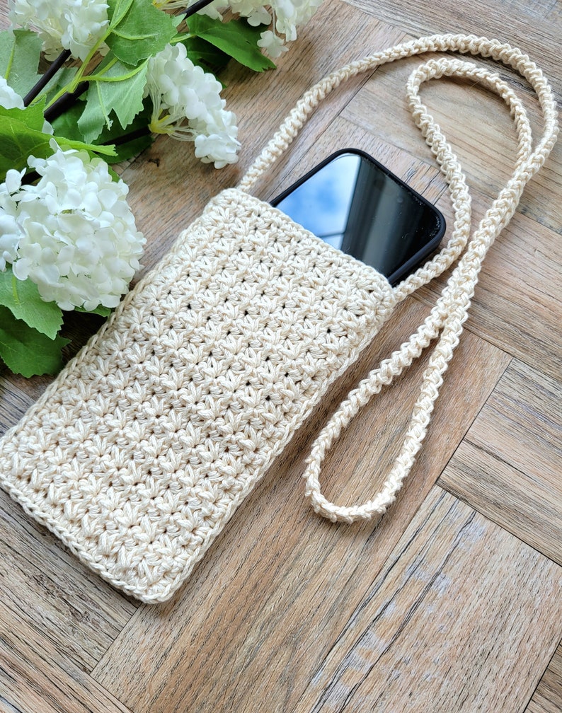 May include: A crocheted beige phone case with a long strap. The case is lying on a wooden surface with a white flower in the background.
