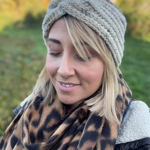 May include: A woman wearing a beige crocheted headband with a knotted design. She is also wearing a leopard print scarf and a black jacket.