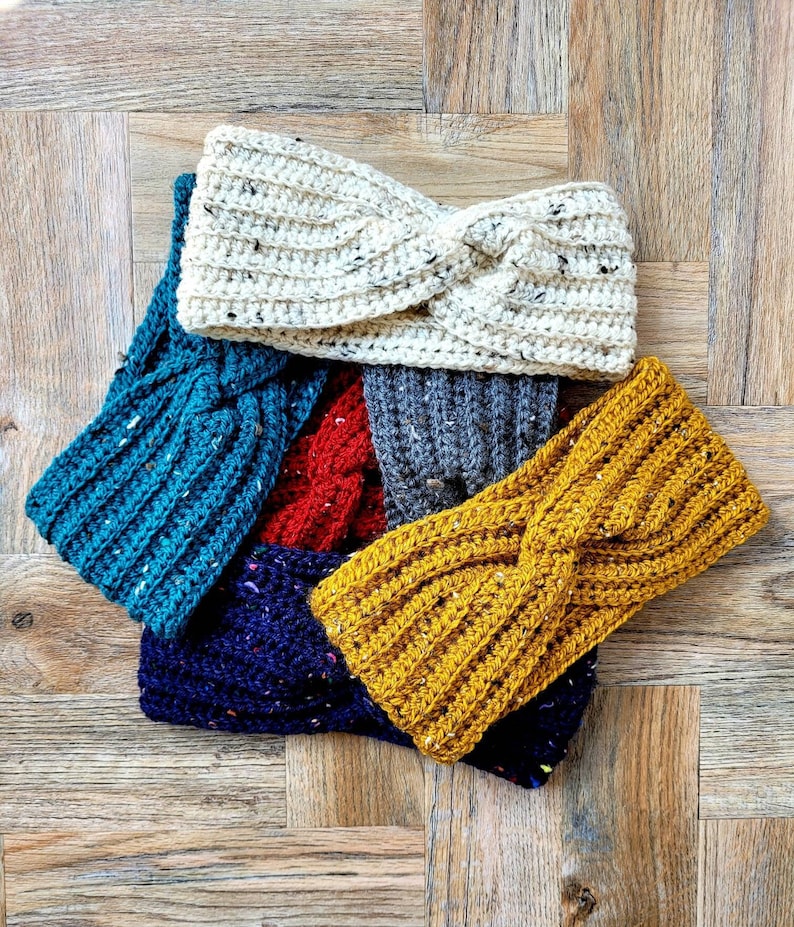 May include: Five crocheted headbands in different colors and patterns. The headbands are arranged in a pile, with the top headband being white with black flecks. The other headbands are teal, red, navy blue with white flecks, and yellow.