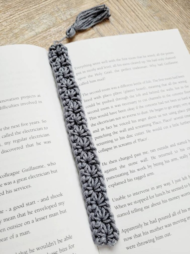May include: A gray crocheted bookmark with a tassel. The bookmark is laying on top of an open book with text about a tradesman.
