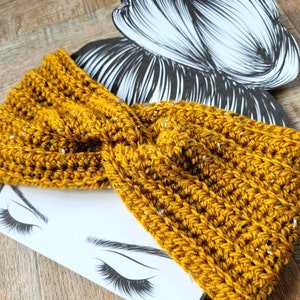 May include: A mustard yellow crocheted headband with a textured pattern. The headband is folded in a twist and is lying on a white surface with a drawing of a woman's eyes and eyelashes.