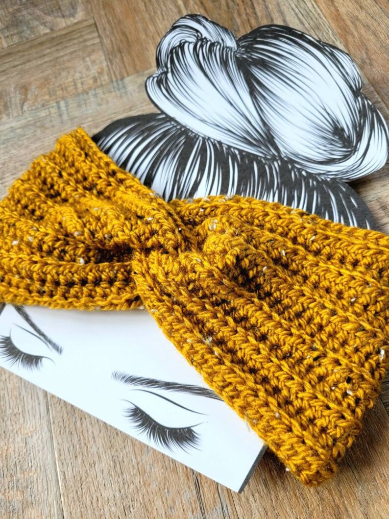 May include: A mustard yellow crocheted headband with a textured pattern. The headband is folded in a twist and is lying on a white surface with a drawing of a woman's eyes and eyelashes.