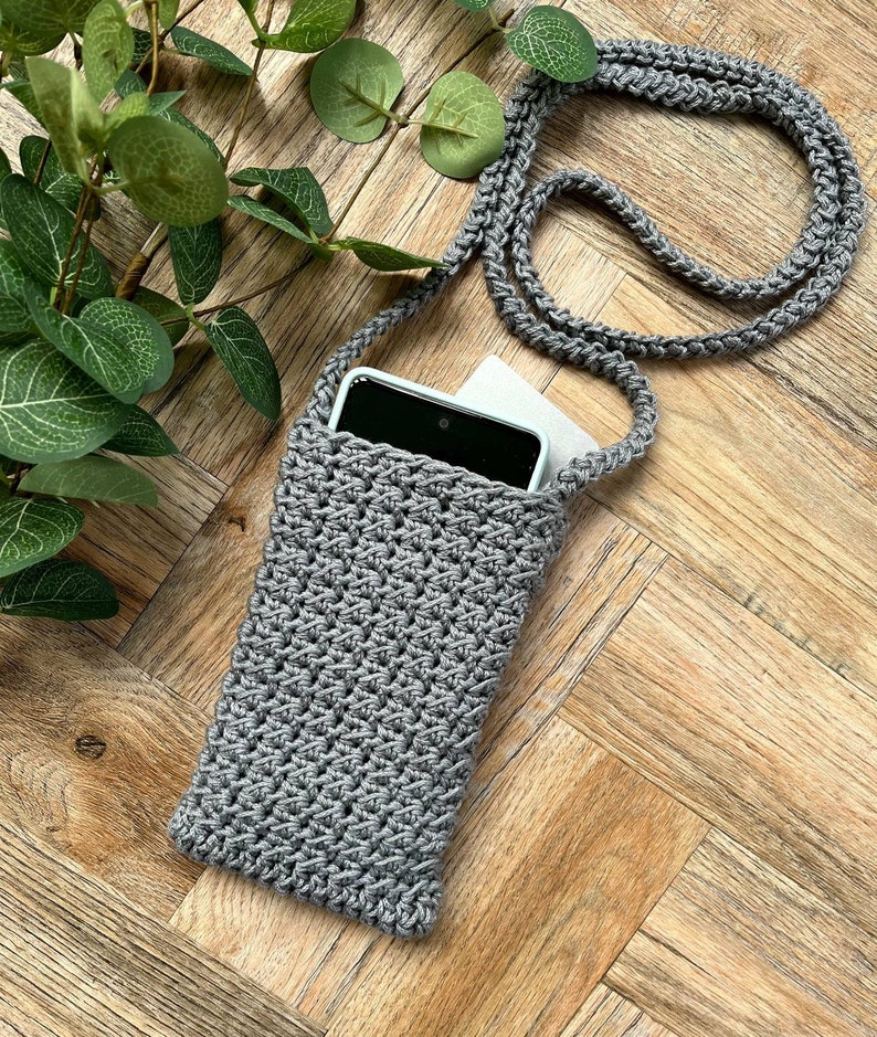 May include: A gray crocheted phone case with a long strap. The case is lying on a wooden surface with a smartphone inside.