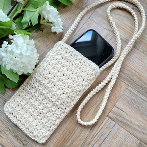 May include: A crocheted beige phone case with a long strap. The case is made with a simple stitch pattern and has a black phone inside.