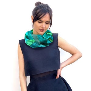 Könnte beinhalten: Eine Frau trägt ein dunkelblaues ärmelloses Oberteil und einen Rock. Ein leuchtend grüner und türkisfarbener Schal mit gewebtem Design ist um den Hals drapiert. Der Schal bildet einen Kontrast zur dunklen Kleidung.