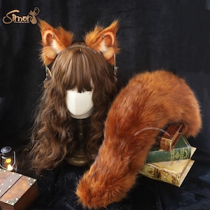 May include: A set of faux fur fox ears and a matching long, bushy fox tail in a vibrant reddish-brown color. The ears are mounted on a headband and feature small metallic charms. The set is displayed with a brown wig on a mannequin head.
