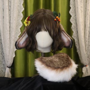 Può includere: Un set di accessori per costumi a tema bosco. Il set include un paio di orecchie da coniglio in finta pelliccia marrone e bianca con una corona floreale e una coda soffice abbinata. Le orecchie sono attaccate a una parrucca. Gli accessori sono esposti su una testa di manichino.