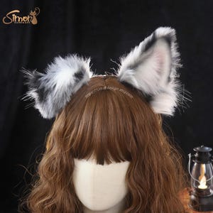 May include: A pair of faux fur animal ears in shades of white, gray, and black, attached to a headband. The ears feature long, fluffy texture with pointed tips and a soft inner ear detail. The text Simon Handmade and simordesign.etsy.com are visible.