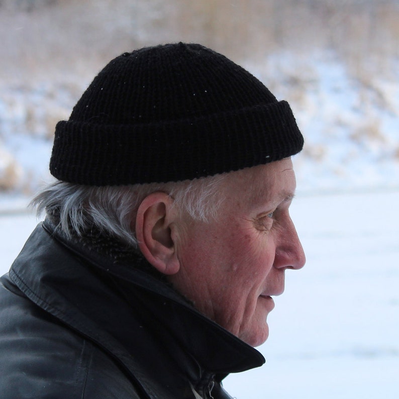 K&ouml;nnte beinhalten: Eine schwarze Strickm&uuml;tze. Die M&uuml;tze wird von einer Person mit grauem Haar und einer schwarzen Jacke getragen. Der Hintergrund ist eine verschneite Szene im Freien.