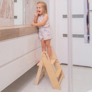 May include: A light-colored wooden step stool with two tiers and side cut-out handles. The stool features a sturdy A-frame design with visible metal hardware connecting the side panels to the steps, providing a stable platform for reaching elevated surfaces.