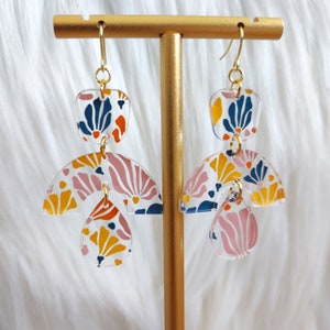 May include: Clear acrylic earrings with a gold-tone hook. The earrings feature a layered design with a square top, a half-circle middle, and a teardrop bottom. The earrings are decorated with a colorful floral pattern in shades of blue, orange, pink, and yellow.