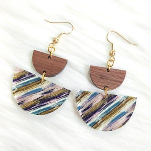 May include: A pair of dangle earrings with a modern design. Each earring features a half-circle of dark wood above a semi-circular pendant with a colorful abstract pattern in shades of blue, purple, pink, and gold. The earrings have gold-colored ear wires.