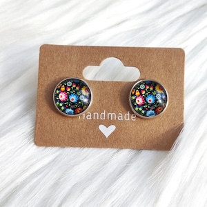 May include: Two round stud earrings with a floral design in shades of pink, blue, and yellow on a black background. The earrings are set in silver-colored metal and displayed on a brown card with the word "handmade".