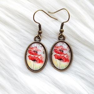 May include: A pair of oval-shaped earrings with a bronze finish. Each earring features a vibrant red poppy flower design set against a soft, off-white background. The earrings have a classic hook closure.