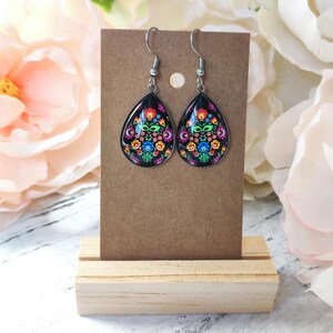 May include: A pair of teardrop-shaped earrings with a black background and a colorful floral design. The earrings are displayed on a brown card, held by a wooden stand. The floral design includes red, blue, green, and yellow flowers.