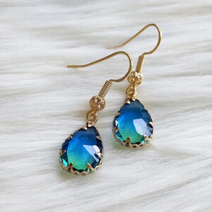 May include: A pair of gold-colored drop earrings with teardrop-shaped blue and green faceted stones. The earrings have a decorative gold-colored setting around the stones and gold-colored fish hook ear wires. The earrings are displayed on a white, fluffy surface.