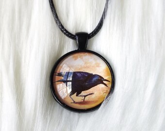 May include: A round pendant necklace featuring a black crow illustration. The crow is set against a warm, textured background within a black metal frame. The necklace has a black cord.