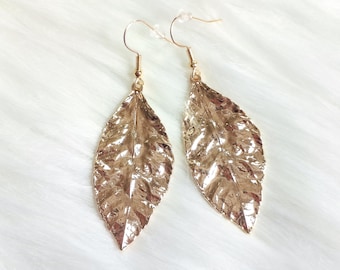 May include: A pair of gold-colored leaf-shaped earrings. The earrings have a textured surface and are suspended from gold-colored ear wires. The earrings are displayed on a white, fluffy surface.