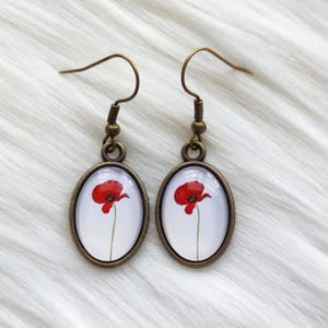 May include: A pair of oval-shaped earrings with a bronze finish. Each earring features a red poppy flower on a white background, encased in a glass dome. The earrings have a hook closure.