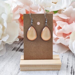 May include: A pair of teardrop-shaped earrings with a light orange center and a dark bronze border. The earrings hang from silver-colored hooks. The earrings are displayed on a brown card, set on a wooden stand, with a floral background.