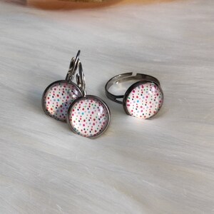 May include: A jewelry set featuring a pair of leverback earrings and an adjustable ring. Each piece has a round, silver-toned setting with a white background and a pattern of colorful polka dots. The set is displayed on a white, fluffy surface.