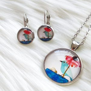 May include: A jewelry set featuring a pendant necklace and matching earrings. Each piece has a round, silver-toned frame with a watercolor design of a blue bird under a red umbrella. The bird is perched on a blue surface.