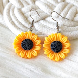 May include: A pair of sunflower earrings. Each earring features a bright yellow sunflower with a black center. The earrings have silver-colored hooks. The background is a white, textured surface.