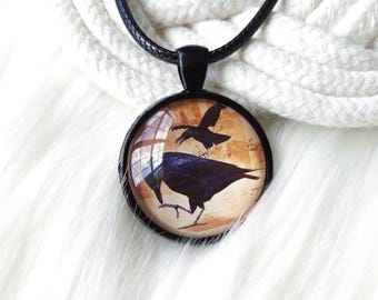 May include: Black pendant necklace featuring a round glass pendant with a raven illustration. The raven is depicted in black against a sepia-toned background. The pendant is set in a black metal frame and hangs from a black cord.