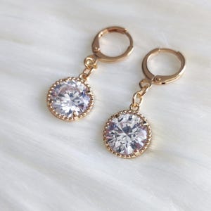 May include: A pair of gold-toned drop earrings featuring circular settings with a beaded edge. Each earring holds a single large, clear, round-cut faceted stone suspended from a hinged hoop closure, creating a classic and reflective jewelry piece.