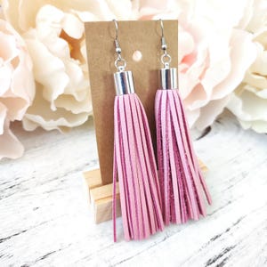 May include: A pair of pink tassel earrings with silver-colored metal accents. The earrings are displayed on a brown card, with a white and pink floral background. The tassels are made of thin strips of pink material.