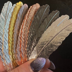May include: A collection of metal feather-shaped bookmarks in various metallic finishes, including silver, gold, rose gold, and gunmetal. The bookmarks are fanned out, showcasing their intricate feather detailing. The image is well-lit, highlighting the different colors and textures.