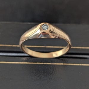 May include: A polished rose gold ring featuring a single, small, round, clear gemstone set flush into the center of the band. The band has a smooth, tapered design that widens slightly at the top to accommodate the circular stone setting.