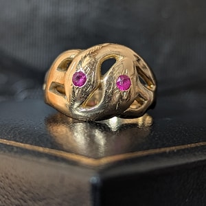 Op de afbeelding: Een gouden ring met een verstrengeld, opengewerkt ontwerp dat twee slangachtige koppen vormt. Elke kop is geaccentueerd met een kleine, ronde, levendige roze-rode edelsteen die in het metalen oppervlak is gezet voor een textuurafwerking.