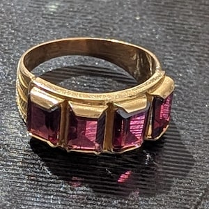 Peut inclure: Une bague dorée ornée de quatre pierres précieuses rectangulaires, d'un violet profond. La bague a une bande épaisse avec un motif texturé sur les côtés. Les pierres sont disposées en rangée.