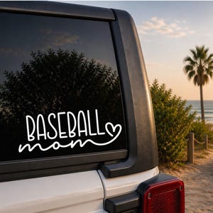 May include: White vinyl decal on a car window. The decal reads "BASEBALL mom" in a handwritten style font, with a heart symbol. The background shows a beach scene with palm trees and the ocean.