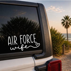 May include: White vinyl decal with the words "AIR FORCE wife" in a handwritten style, with a heart symbol. The decal is on the rear window of a white vehicle, with a beach scene in the background.