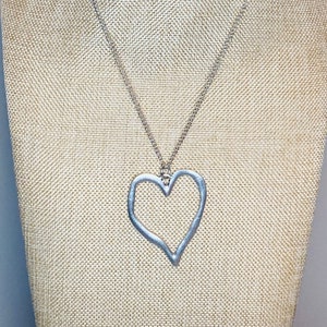 May include: A silver necklace with a large, open heart pendant. The heart is made of a thin, textured metal and has a slightly irregular shape.