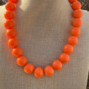 May include: A bright orange chunky necklace made of large round beads. The necklace is displayed on a tan fabric mannequin.