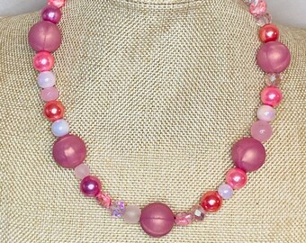 May include: A pink and white beaded necklace with large, round beads. The necklace is made of various shades of pink and white beads, including some with a shimmery finish.