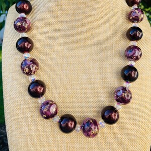May include: A purple and gold glass bead necklace with clear crystal accents. The necklace is on a brown fabric mannequin.