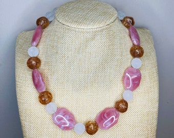 May include: A pink and white gemstone necklace with brown beads. The necklace is on a beige fabric mannequin.
