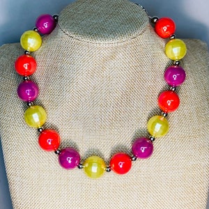 Puede incluir: Un collar colorido hecho de cuentas grandes y redondas en tonos amarillos, naranjas y morados. Las cuentas están enhebradas en una cadena plateada.