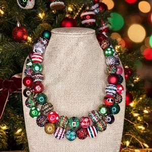 May include: A double-strand chunky beaded necklace featuring a variety of round beads in red, green, white, and gold colors. The beads include solid, striped, polka-dot, and glitter-covered patterns, arranged on a beige fabric display bust.