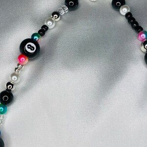 May include: A beaded necklace with a black 8-ball charm, black, white, silver, and iridescent beads. The necklace has a silver clasp and is displayed on a white satin background. The beads are various sizes and shapes.